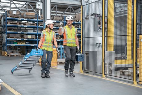 Zwei Arbeiter in Warnweste, Arbeitskleidung und Schutzhelm laufen durch die Produktion. Der linke Arbeiter zieht eine Multimaster 5 auf Rollen hinterher.